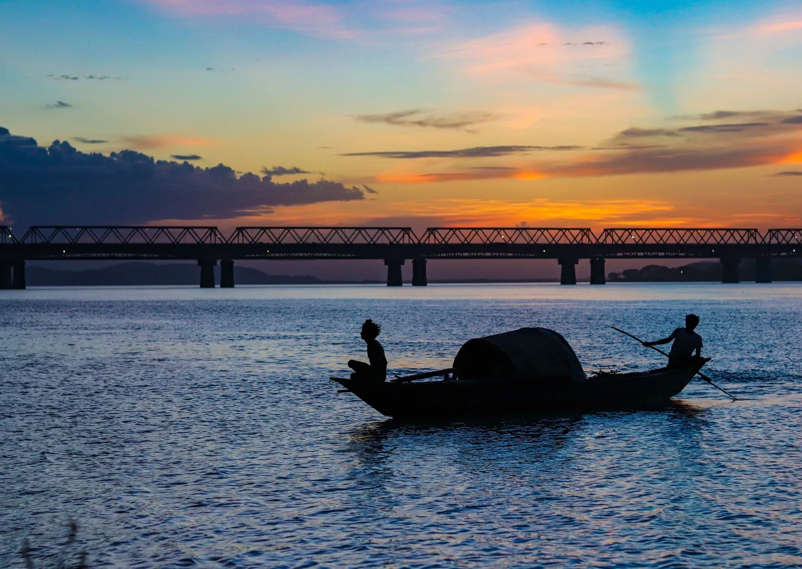 Fishermen in a country boat on the Brahmaputra at sunset, a long bridge behind them