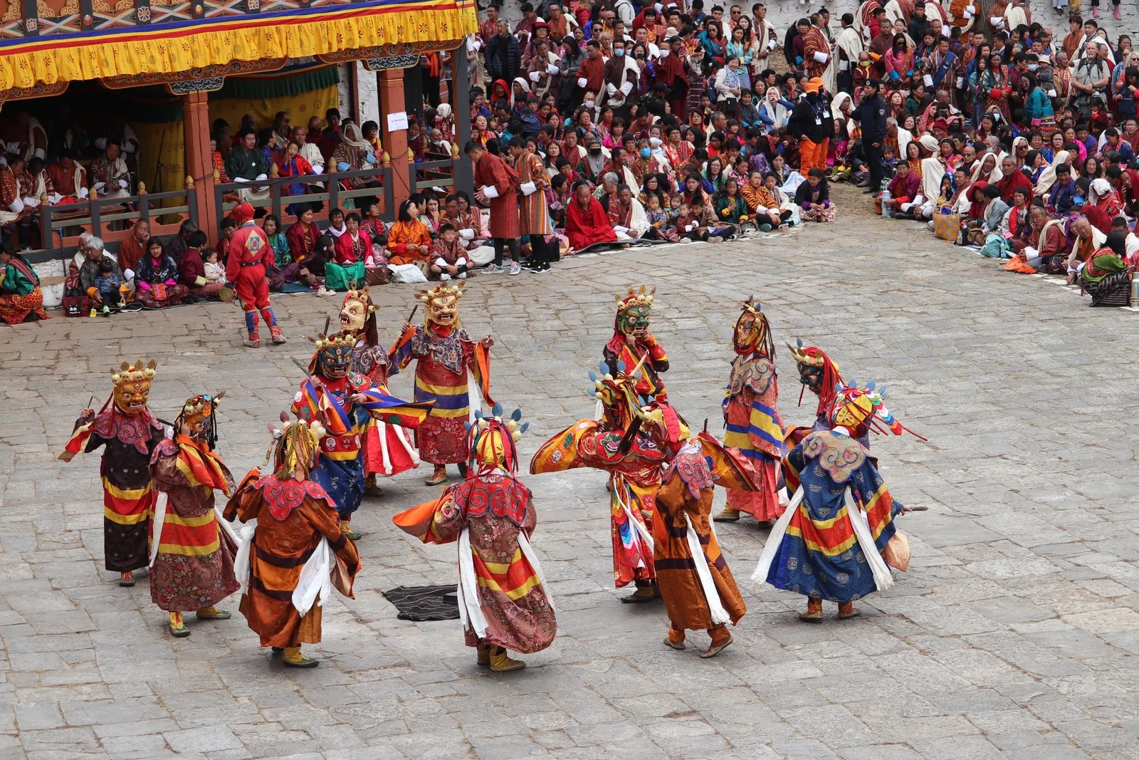Masked dancers turning in a dzong courtyard at a tsechu, the crowd watching, Bhutan
