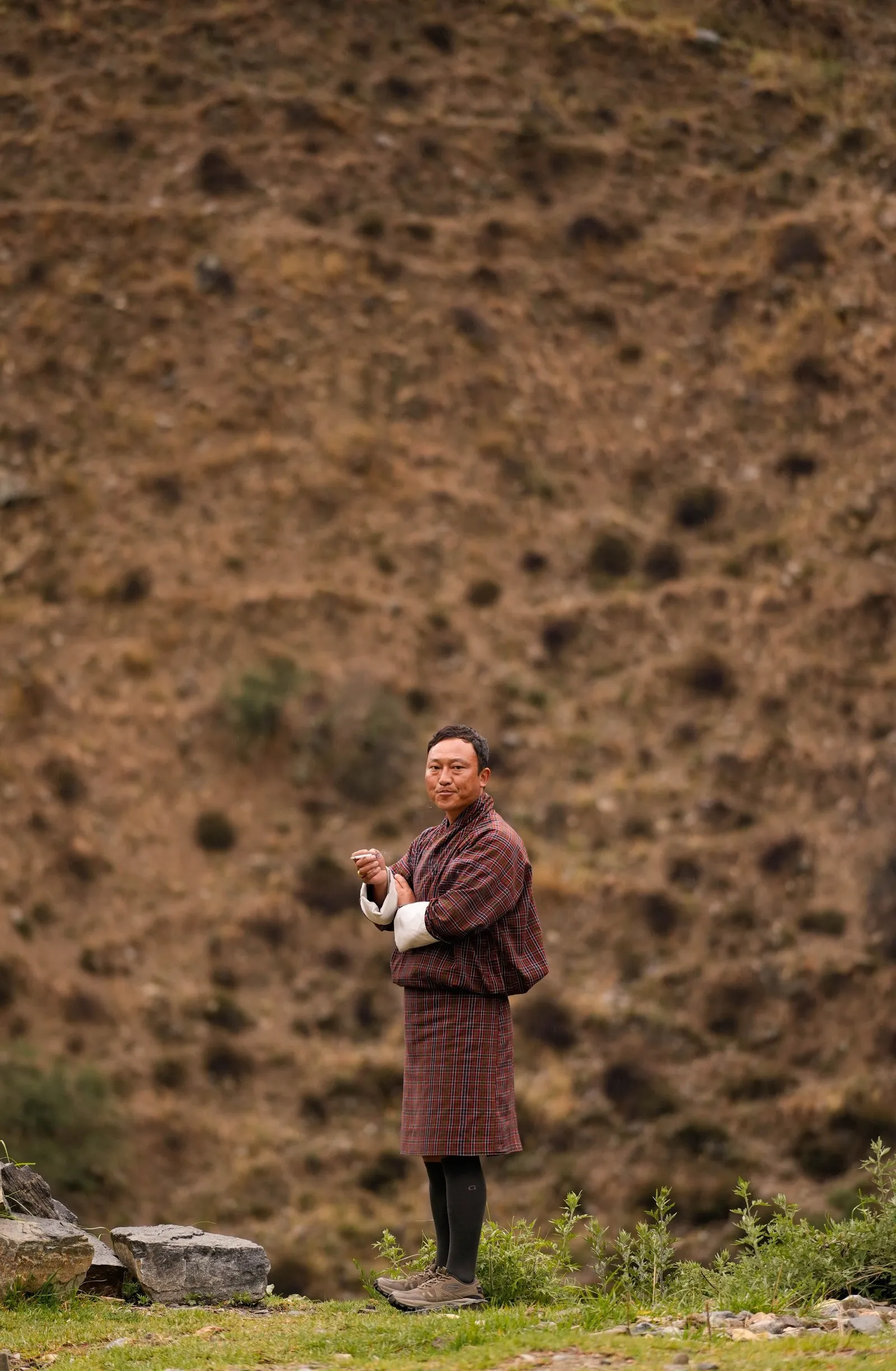 A man in a gho standing on a grassy hillside, Bhutan