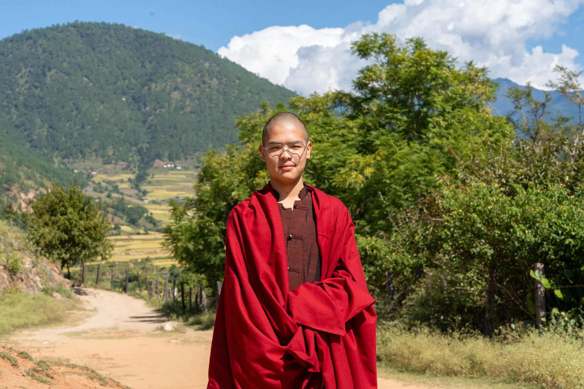 A young monk in a red robe on a farm track, terraced fields and forested hills behind, Bhutan