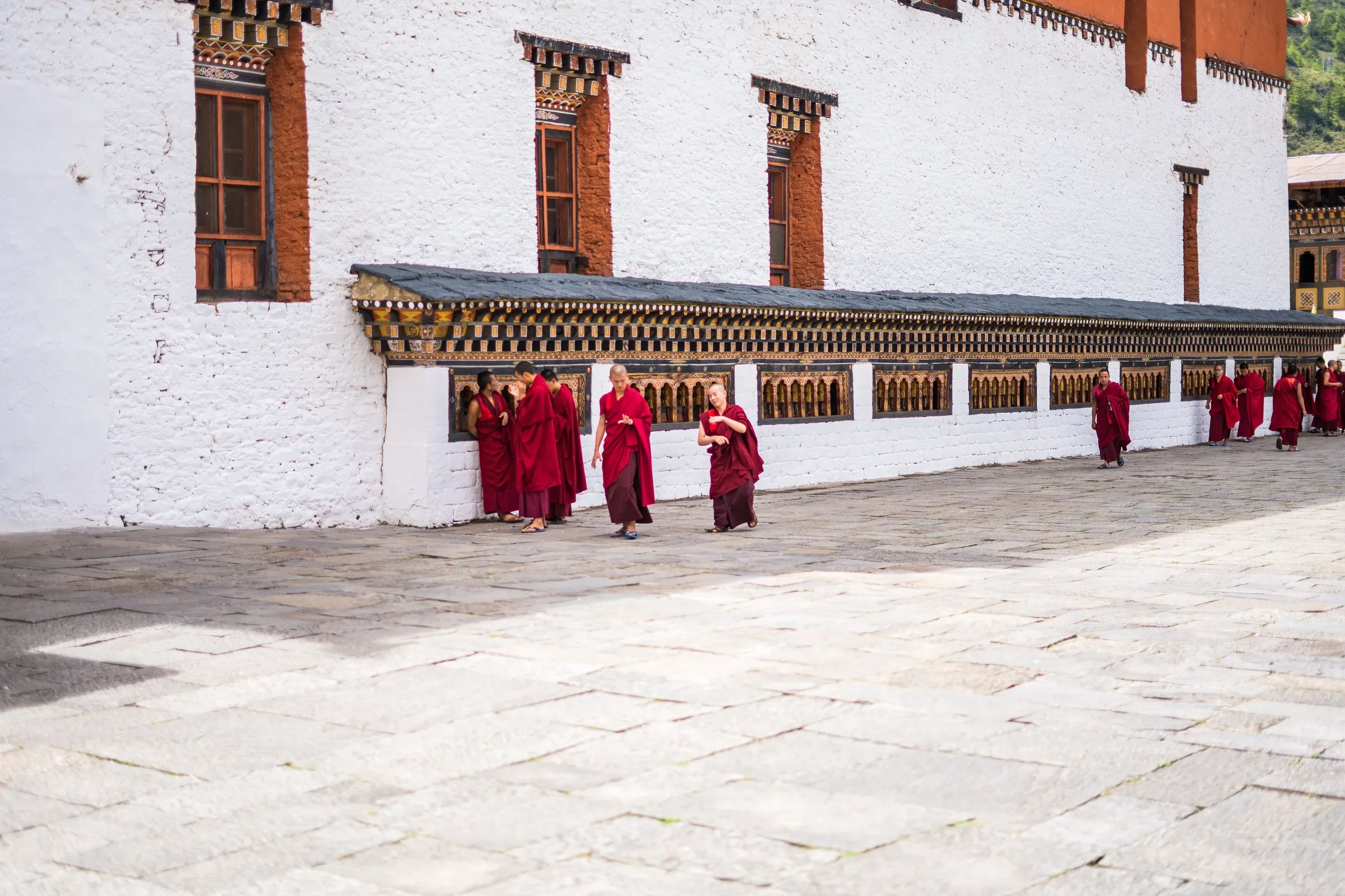Monks walking past a long wall of prayer wheels in a dzong courtyard, Bhutan