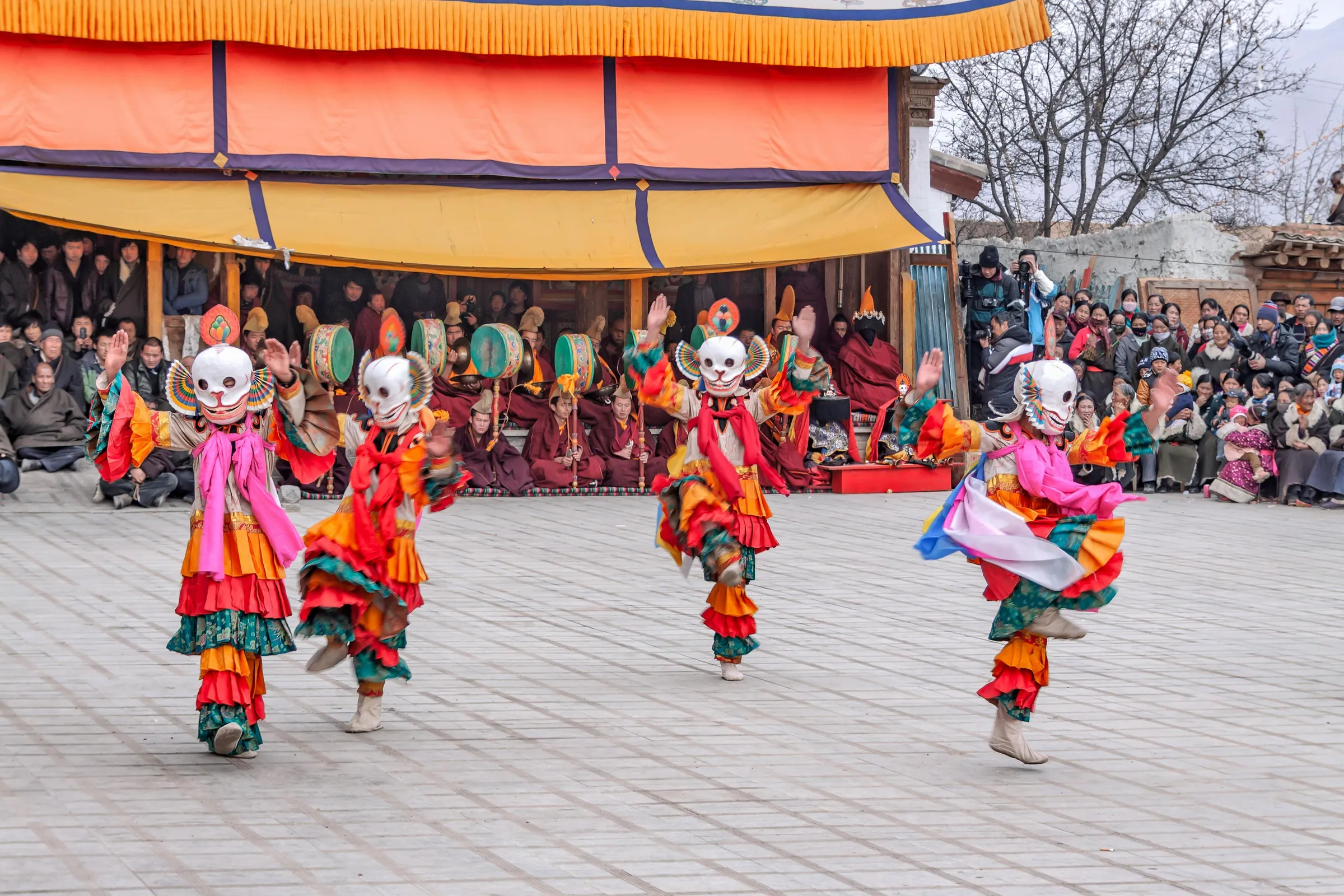 Masked Cham dancers turning in the courtyard of a Ladakh monastery, the crowd watching from the steps