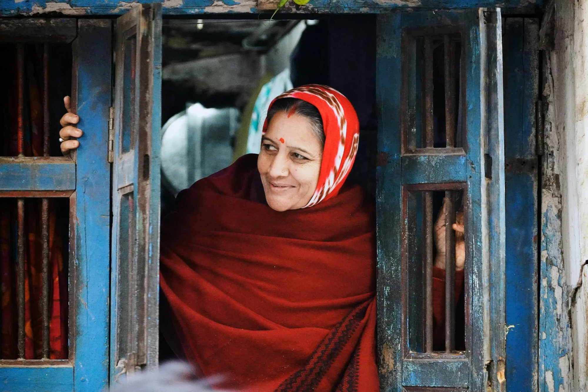A woman in a red shawl smiling from a blue-painted doorway