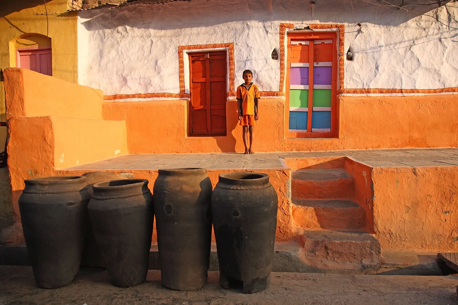 A boy on the porch of a painted house in Hampi village, beside tall clay pots