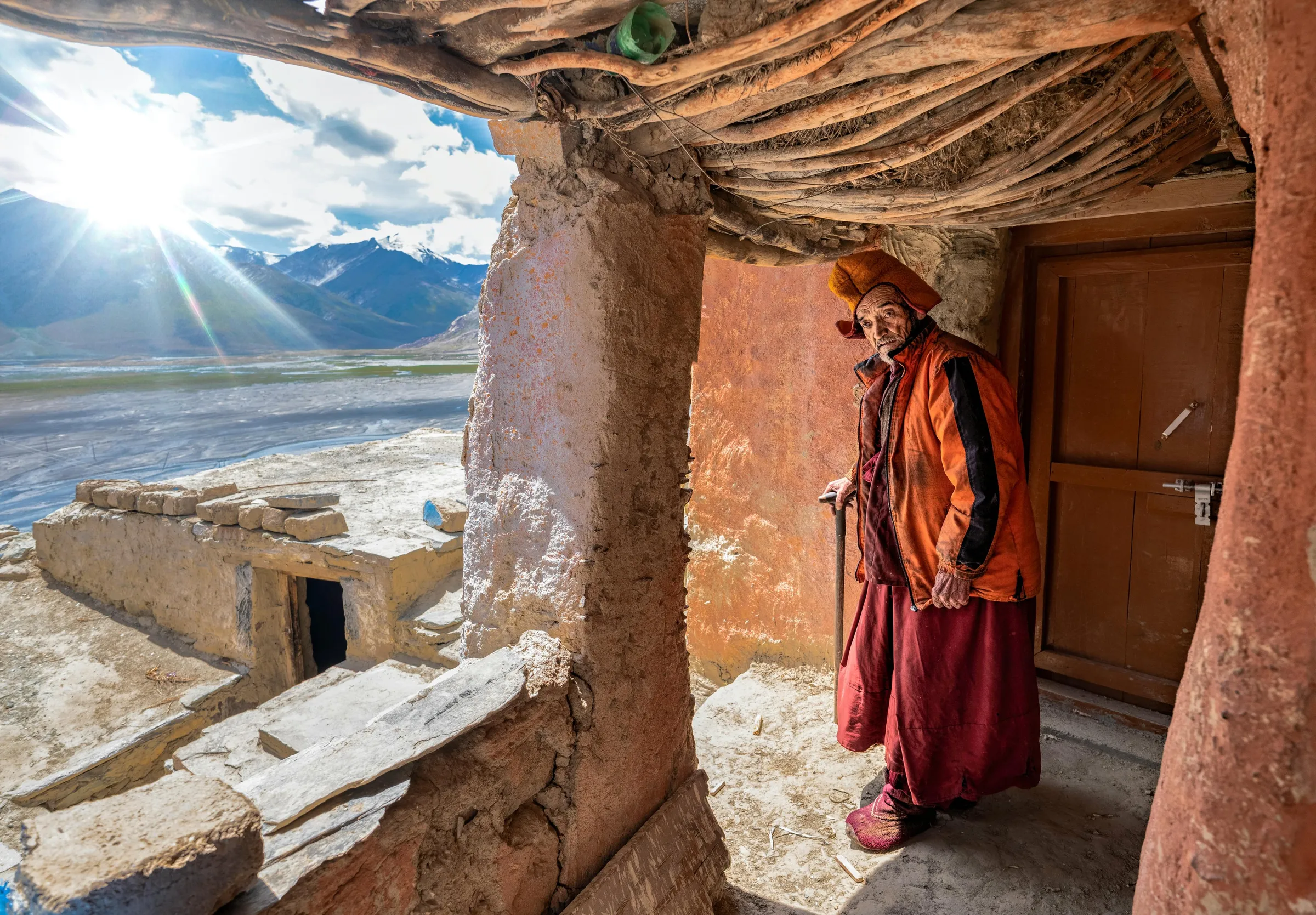 A monk in a red robe at a doorway above a sunlit Ladakh valley