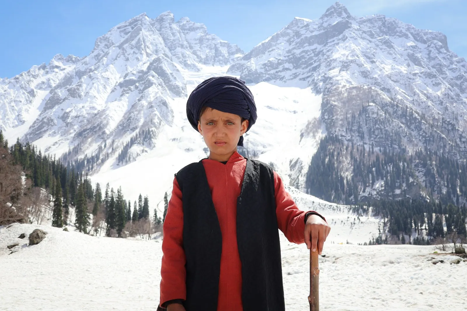 A boy in a black waistcoat and turban with his stick, the snows of Sonamarg behind