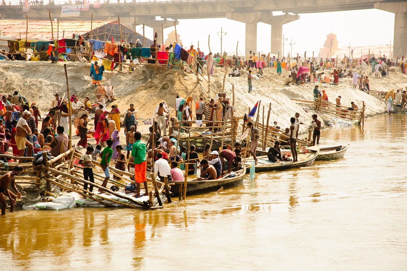 Pilgrims climbing into wooden boats at the riverbank below a bridge, Prayagraj