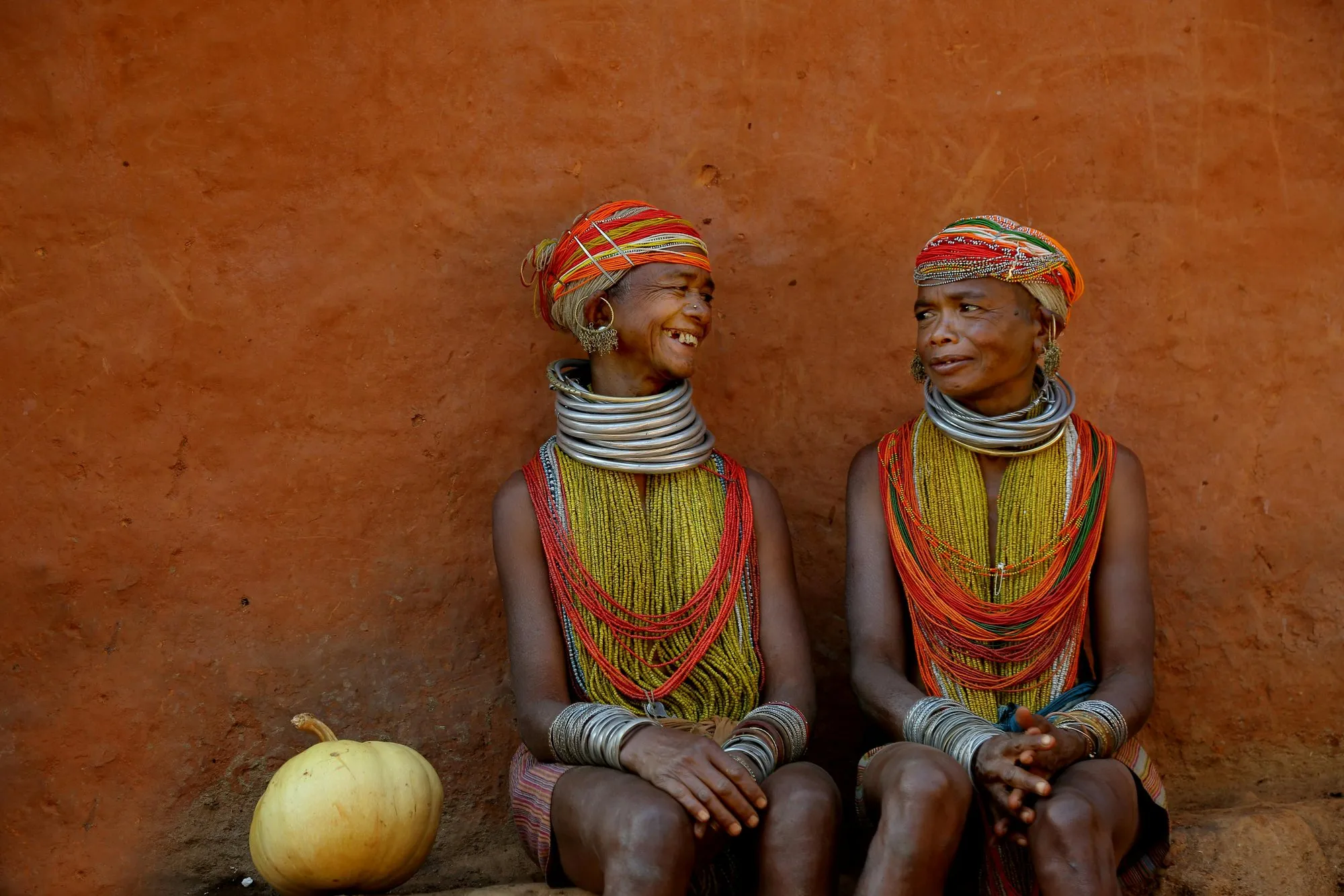 Two women in bead collars and brass neck rings, seated against an ochre wall