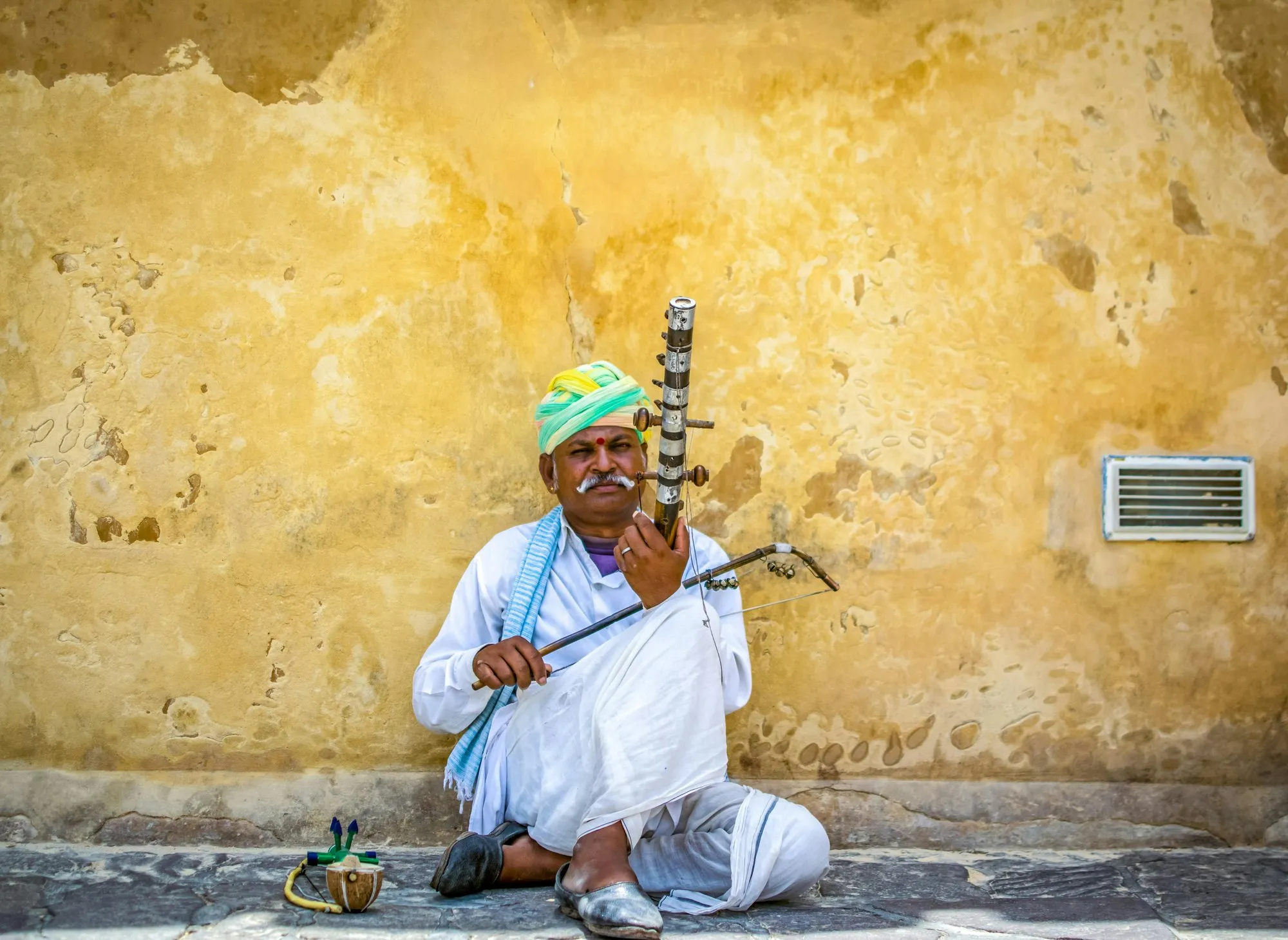 A musician in a green turban playing the ravanahatha against a yellow wall