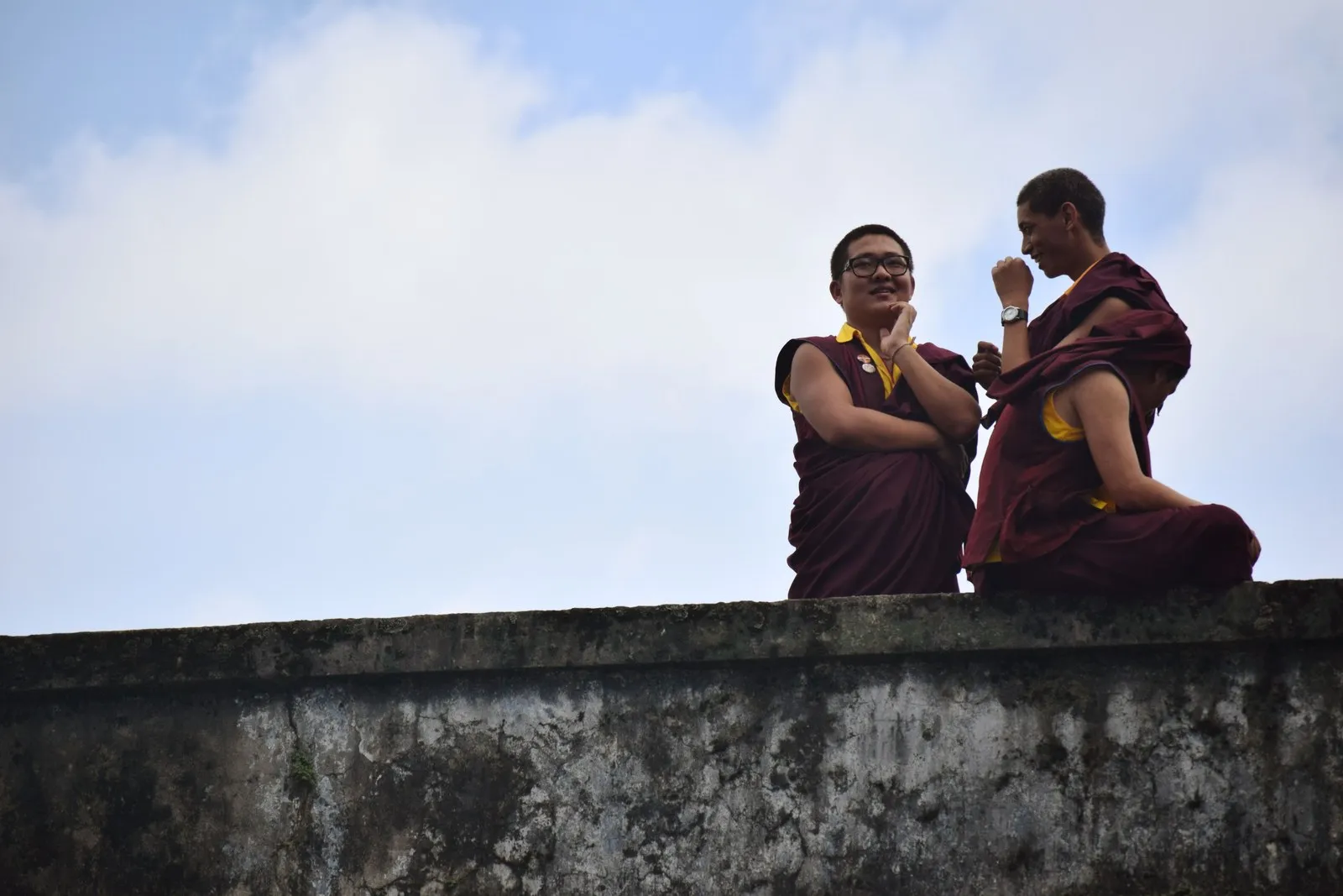 Two monks in maroon robes laughing together on a wall in Kalimpong