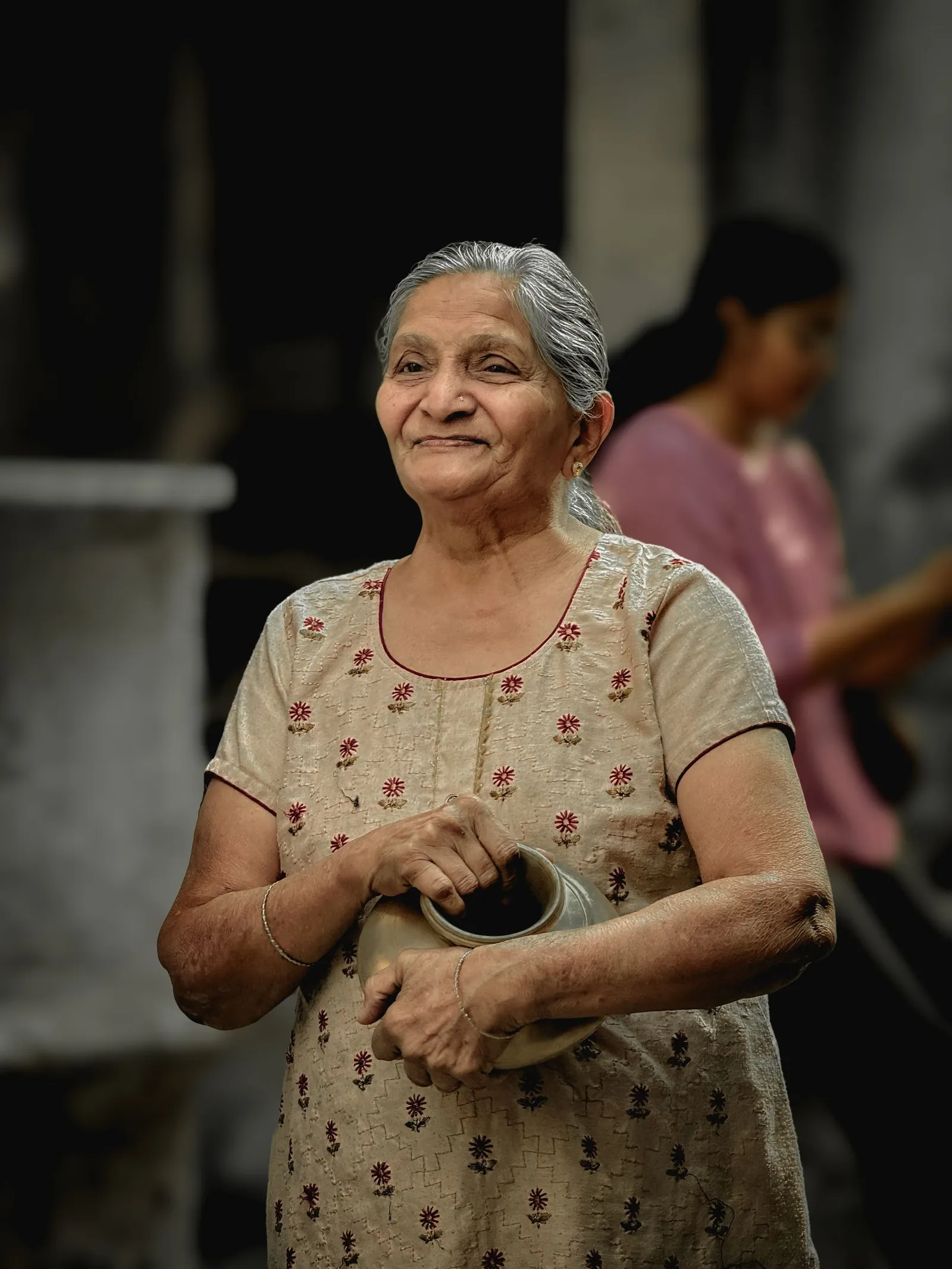An elderly woman smiling, a brass pot in her hands, in Ahmedabad