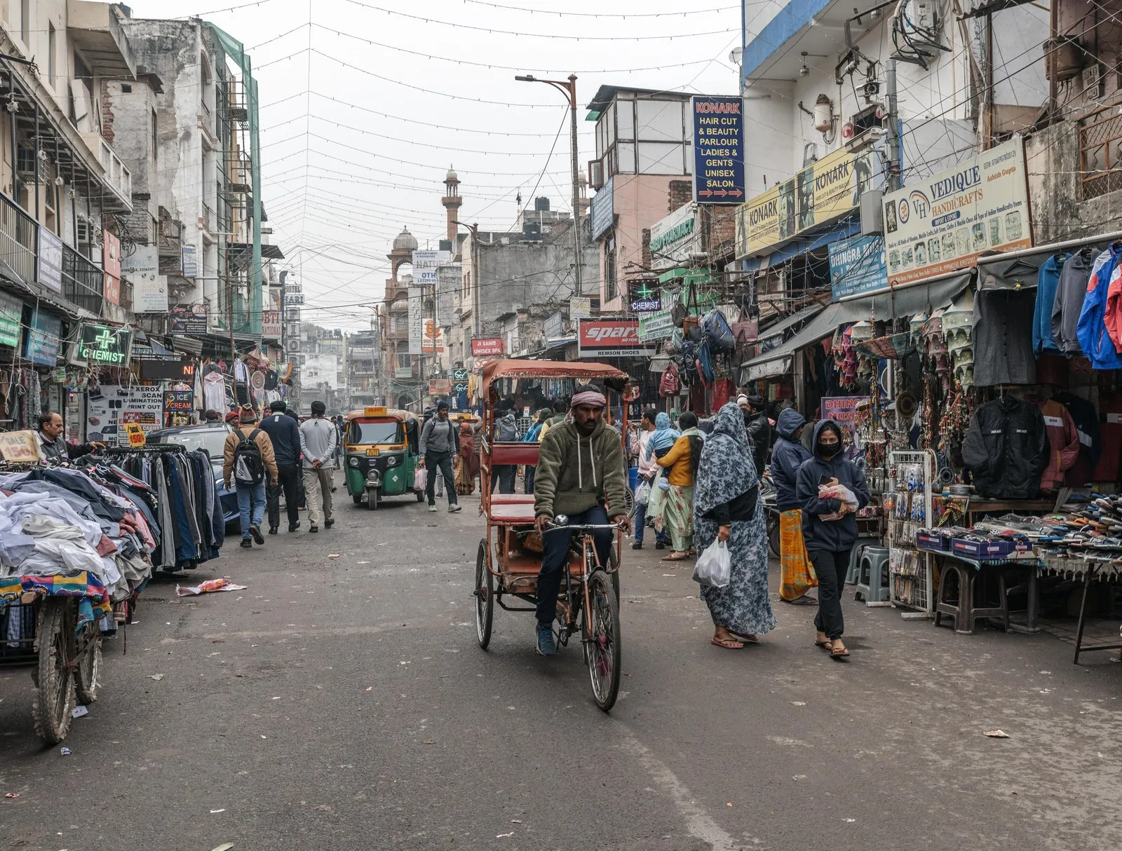 A cycle-rickshaw and shoppers in a market street in Delhi
