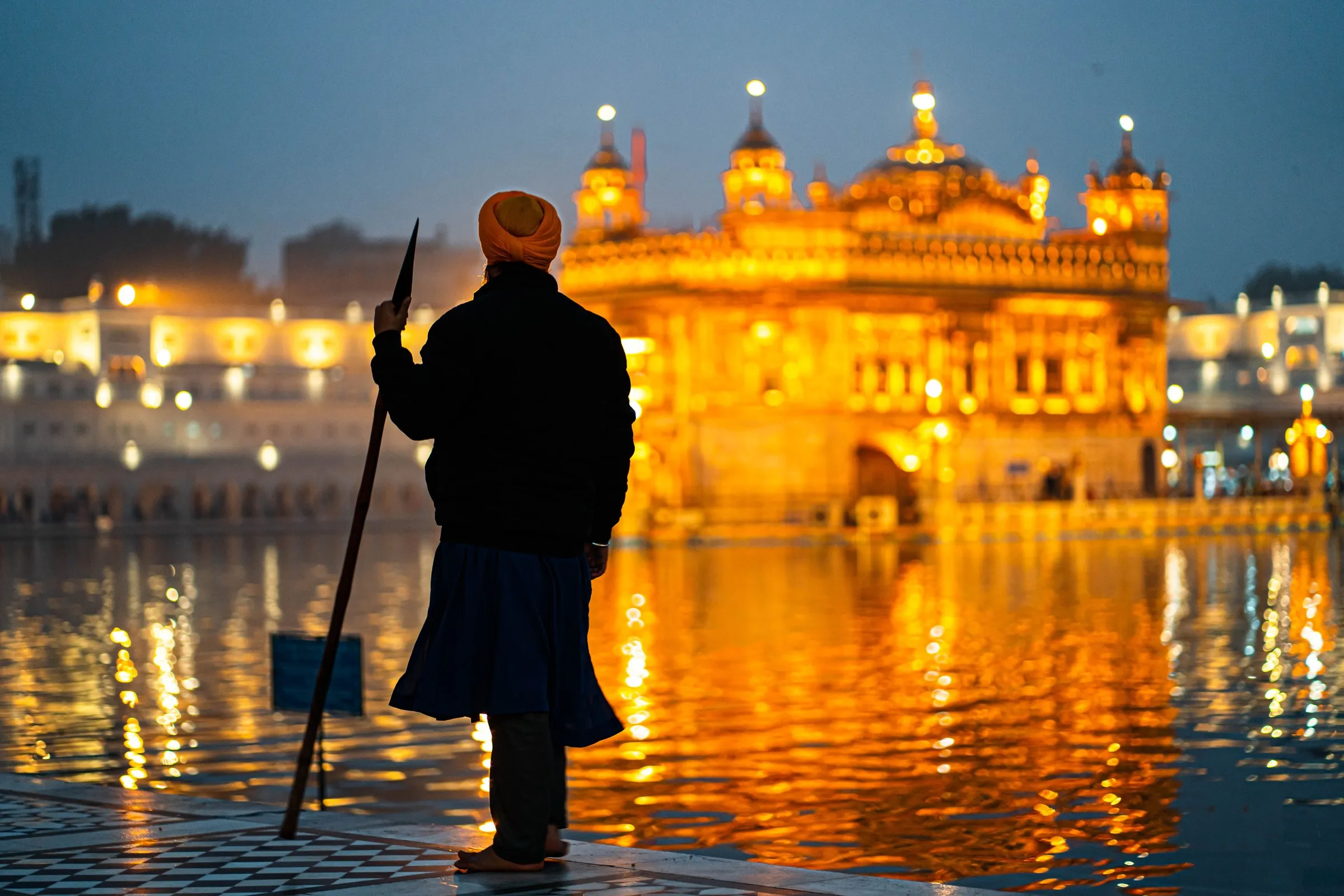 A Sikh sevadar with a spear beside the sacred pool at dusk, the Golden Temple lit beyond
