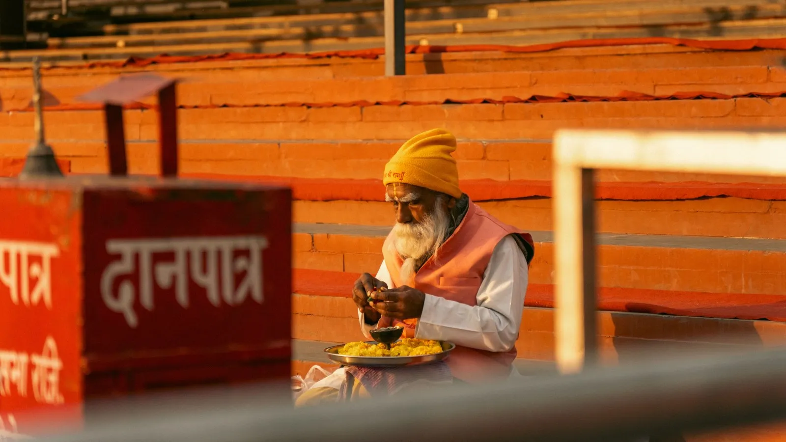 A sadhu seated behind an offering box on a Rishikesh ghat at sunset